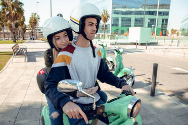 Junges Paar mit Motorradhelmen sitzt zusammen auf einem grünen Fahrrad und spricht auf der Straße - ADSF07819