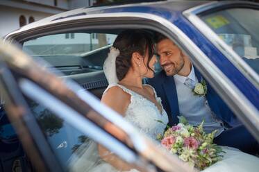 Glückliche Braut im Hochzeitskleid und Bräutigam in stilvollem Anzug sitzen in Oldtimer-Auto und schauen sich gegenseitig an - ADSF07842