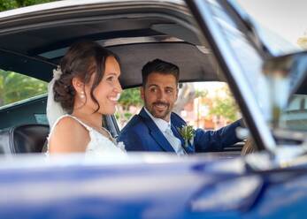 Glückliche Braut im Hochzeitskleid und Bräutigam in stilvollem Anzug sitzen in Oldtimer-Auto und schauen sich gegenseitig an - ADSF07844