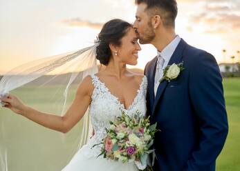 Fröhlicher Bräutigam im eleganten Smoking, der die Braut im Hochzeitskleid hochhebt, während er abends an einer tropischen grünen Küste steht - ADSF07845