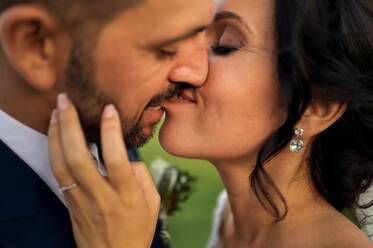 Seitenansicht des fröhlichen Bräutigams im eleganten Smoking, der die Braut im Hochzeitskleid hochhebt, während er am Abend an einer tropischen grünen Küste steht - ADSF07847