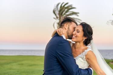 Seitenansicht des fröhlichen Bräutigams im eleganten Smoking, der die Braut im Hochzeitskleid hochhebt, während er am Abend an einer tropischen grünen Küste steht - ADSF07851