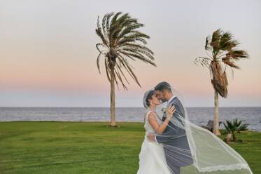 Side view of cheerful groom in elegant tuxedo lifting bride in wedding gown while standing on tropical green coast in evening time - ADSF07852