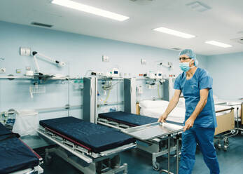 Sanitäter in blauer Uniform und Schutzmaske, der ein Tablett auf einen Wagen im Krankenhauszimmer neben leeren Betten stellt - ADSF07955