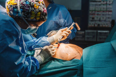 Seitenansicht eines ernsten jungen Arztes mit Schutzmaske und Kappe, der eine Operation mit Instrumenten und einer Krankenschwester durchführt - ADSF07960