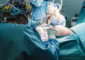 Side view of serious young doctor in protective mask and cap making surgery with instruments and crop nurse - ADSF07961