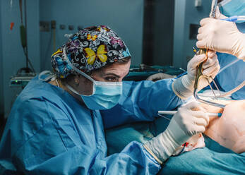 Side view of serious young doctor in protective mask and cap making surgery with instruments and crop nurse - ADSF07963