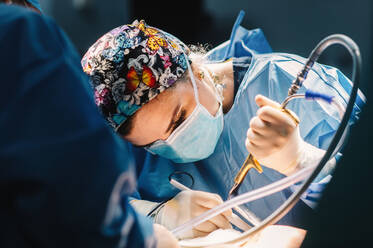 Side view of serious young doctor in protective mask and cap making surgery with instruments and crop nurse - ADSF07970