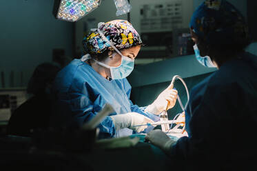 Side view of serious young doctor in protective mask and cap making surgery with instruments and crop nurse - ADSF07971