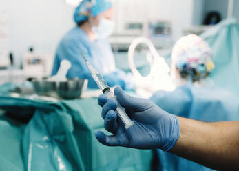 Die behandschuhte Hand mit der vorbereiteten Injektion und die unscharfen Chirurgen bei der Arbeit im Operationssaal - ADSF07972