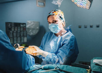 Doctor in surgical gown holding prepared silicone implant for mammoplasty and crop nurse assisting during operation - ADSF07973
