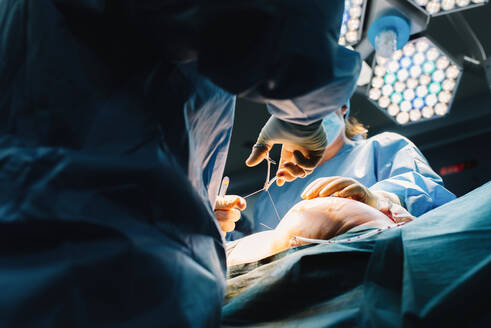 Von unten: Plastischer Chirurg näht die Brust einer Patientin nach dem Einsetzen von Implantaten im Operationssaal zu - ADSF07974
