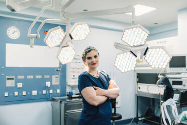 Adult woman in medical uniform crossing arms and looking at camera while standing near lamps in modern operating theater - ADSF07978
