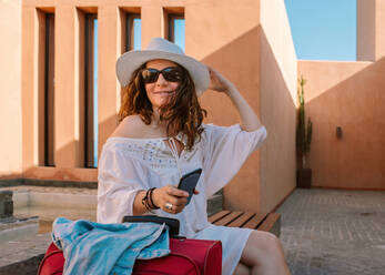 Glückliche erwachsene Frau mit Sonnenbrille und lässigem weißen Kleid, die wegschaut und ihren Hut berührt, während sie allein auf einer Bank neben einem roten Koffer in der Nähe eines Gebäudes bei schönem Sommerwetter sitzt und wartet - ADSF07980