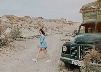 Seitenansicht einer Frau in Shorts und Jeanshemd, die an einem grünen Lastwagen mit Holzaufbau vorbeigeht und in die Ferne auf einen natürlichen Hintergrund schaut - ADSF07986