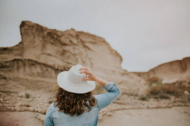 Back view of unrecognizable woman in casual clothes holding hat in her head while walking on desert sandy dunes on gloomy day - ADSF07990