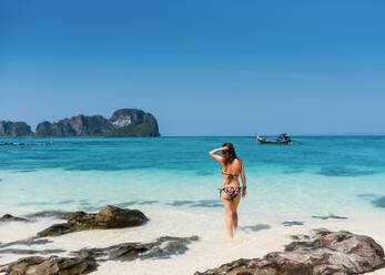 Rückenansicht einer schlanken Touristin im Badeanzug an der Sandküste des türkisfarbenen Ozeans unter blauem Himmel in Thailand - ADSF07997