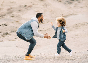 Cheerful bearded ethnic man dressed in stylish clothes playing with curly happy ethnic toddler on bright sandy hills at daytime - ADSF08003