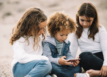 Konzentrierte Kinder in Freizeitkleidung, die sich auf dem Sandboden ausruhen und gemeinsam ein Handy benutzen, während sie tagsüber ein Video ansehen - ADSF08004