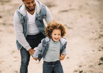 Cheerful bearded ethnic man dressed in stylish clothes holding playing with curly happy ethnic toddler on bright sandy hills at daytime - ADSF08008