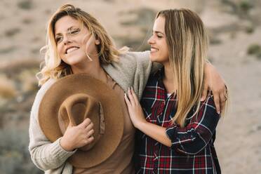 Woman in hat wearing casual clothes tenderly hugging female with long straight hair dressed in check shirt smiling on nature - ADSF08019