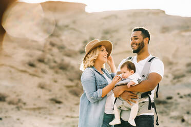 Glücklicher schwarzer bärtiger Mann, der ein kleines Baby trägt und die Hand seiner blonden Frau hält, die Jeans und Hut trägt, während sie auf sandigem Boden steht - ADSF08025