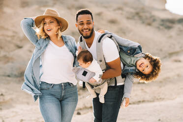 Blond casual woman in hat laughing while black bearded smiling husband carrying children on nature at daytime - ADSF08026