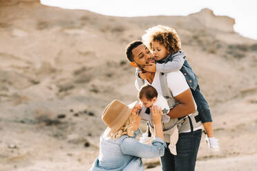 Blond casual woman in hat laughing while black bearded smiling husband carrying children on nature at daytime - ADSF08027