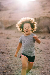 Cheerful active child with curly brown hair dressed in sportive clothes jogging in morning on sandy landscape in backlit - ADSF08033