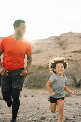 Afroamerikanischer bärtiger aktiver Vater in rotem T-Shirt, der mit einem fröhlichen lockigen Kind durch eine Wüstenlandschaft im Gegenlicht läuft - ADSF08036