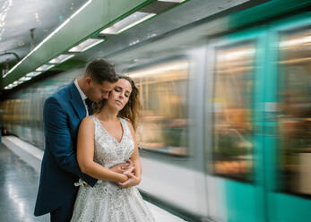 Junge Braut in weißem Hochzeitskleid stehend mit geschlossenen Augen, während Bräutigam stehend hinter und umarmen mit beweglichen Zug auf unscharfen Hintergrund in der U-Bahn - ADSF08046
