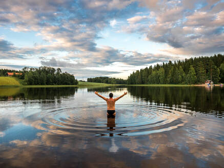 Rückenansicht eines Mannes, der im See steht und die Arme ausbreitet, während er die Landschaft mit Wald und Häusern in Finnland genießt - ADSF08061