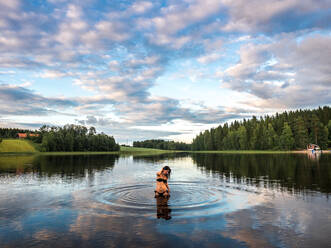 Woman standing in lake enjoying landscape with forest and houses in Finland - ADSF08062
