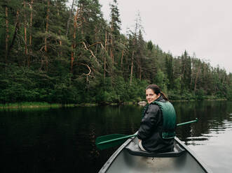 Young smiling woman looking back while boating on forest river in Finland - ADSF08065