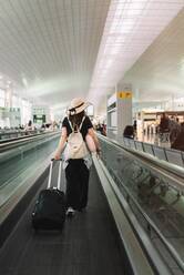 Rückenansicht einer Frau in Freizeitkleidung und Hut mit Gepäck, die auf einer Rolltreppe im Flughafen läuft - ADSF08104