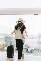 Rückenansicht einer Dame in Freizeitkleidung und Hut mit Gepäck, die auf den Abflug im Flughafen wartet und aus dem Fenster schaut - ADSF08105