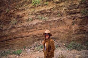 Side view of modern female tourist in stylish outfit with hat and sunglasses looking at camera standing in sandstone ravine and enjoying amazing view of rocky formations - ADSF08108