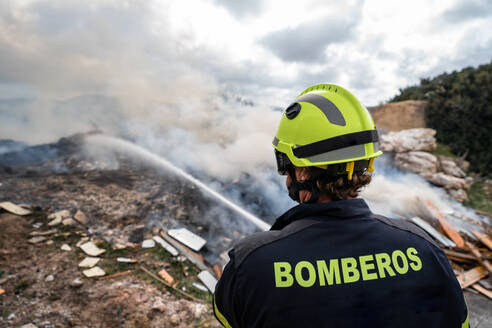 Rückansicht eines tapferen Feuerwehrmanns in Schutzkleidung, der mit einem Schlauch steht und ein Feuer auf einer Müllkippe in den Bergen löscht - ADSF08116