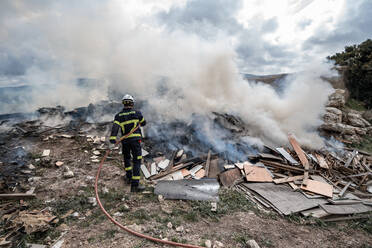 Rückansicht eines tapferen Feuerwehrmanns in Schutzkleidung, der mit einem Schlauch steht und ein Feuer auf einer Müllkippe in den Bergen löscht - ADSF08117