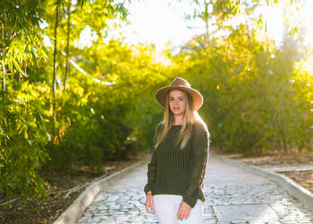 Positive young female in stylish outfit and hat looking at camera while standing on stone pathway in green park in sunny day - ADSF08127