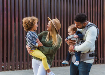 Cheerful stylish young multiracial couple with infant son and little daughter in casual wear walking together along wooden fence on city street - ADSF08131