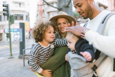 Positive stylish young multiracial parents and cute little daughter stroking head of baby boy carried by father while standing together on city street - ADSF08134