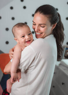 Seitenansicht einer zufriedenen jungen Mutter in Freizeitkleidung lachend mit einem zufriedenen Baby im Arm an einem sonnigen Morgen - ADSF08136