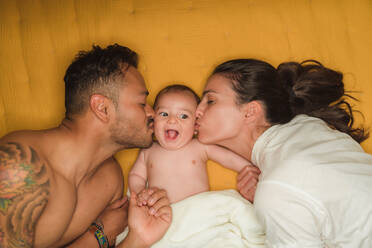 Top view of young happy parents kissing in cheeks little satisfied baby while lying on bed - ADSF08140