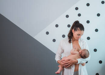 Seriöse schwarzhaarige Mutter in weißem Hemd steht und stillt kleines Baby vor grauer Wand - ADSF08142