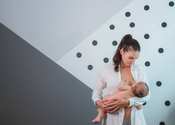 Serious black haired mother in white shirt standing and breastfeeding little baby against gray wall - ADSF08142