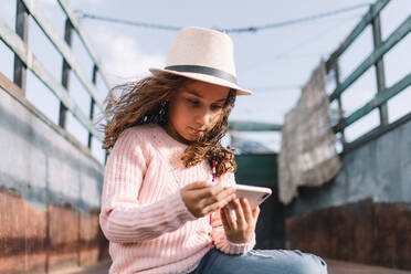 Concentrated girl in straw hat rose knitted jumper and jeans sitting in truck body and browsing mobile phone at windy weather - ADSF08147