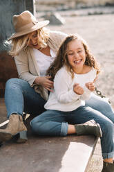 Content woman with blond hair in hat cuddling and tickling daughter in casual clothes while having rest and sitting in old truck body - ADSF08151