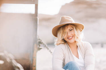 Smiling female in hat with blond hair dressed in bright casual clothes looking at camera sitting near truck body in village at daytime - ADSF08152