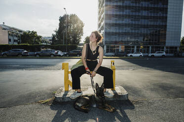 Frau mit Hund sitzt auf einer Bank in der Stadt an einem sonnigen Tag - MEUF01710